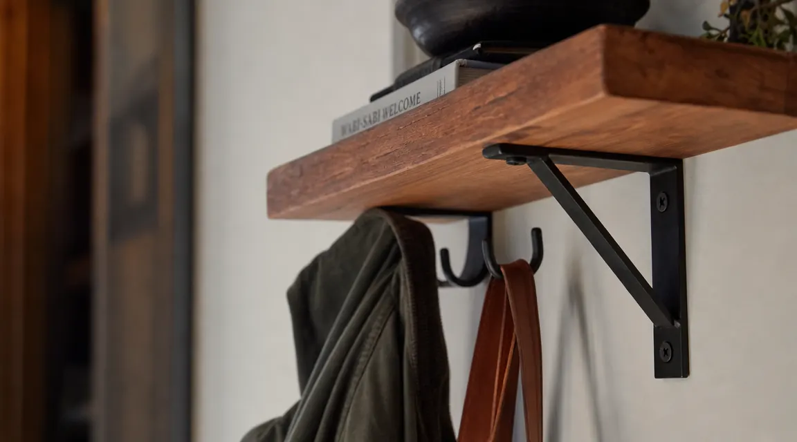 Close-up of a wall-mounted wood and black metal entryway shelf with double hooks holding jackets and bags, showing the wall anchor hardware visible at the bracket